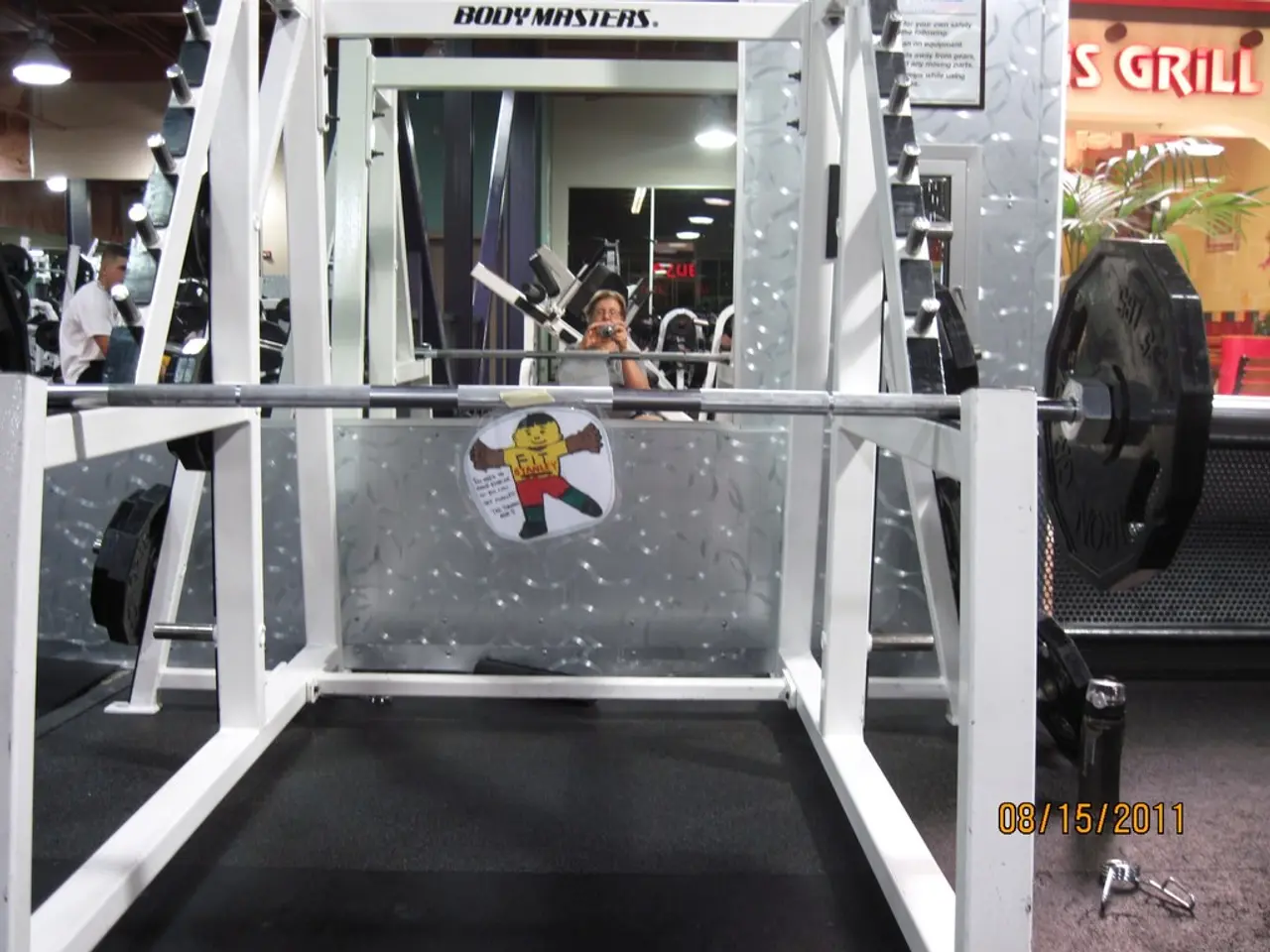 This image is clicked in a gym. In the front, there is a stand alone with dead weights and rod. At...