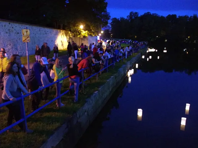 Here we can see candle lights in a box on the water. On the left there are few people standing on...